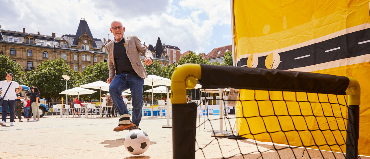 Verkehrsminister Winfried Hermann spielt Fußball mit Gewichten an den Füßen.
