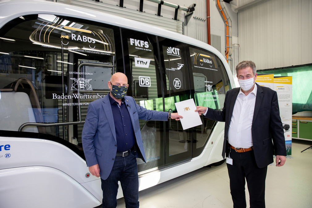 Verkehrsminister Winfried Hermann und Prof. Dr. Hans-Christian Reuss (FKFS) mit Übergabemappe vor dem autonomen Shuttlebus