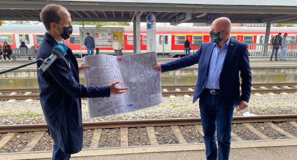 Verkehrsminister Winfried Hermann und der Oberbürgermeister von Crailsheim, Dr. Christoph Grimmer stehen am Crailsheimer Bahnhof und halten gemeinsam einen Bauplan, den sie betrachten