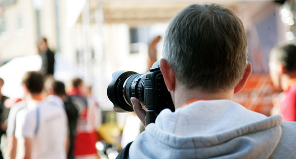 Ein professionel aussehender Fotograf blickt durch seine Kamera und macht ein Foto von mehreren Menschen in einiger Entfernung.