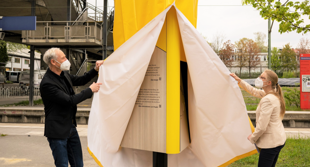 Ein Mann und eine Frau enthüllen eine Informationssäule am Bahnhof in Konstanz.