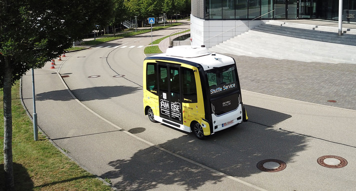 Ein kleines Busshuttle fährt auf einer Straße in eine Kurve.