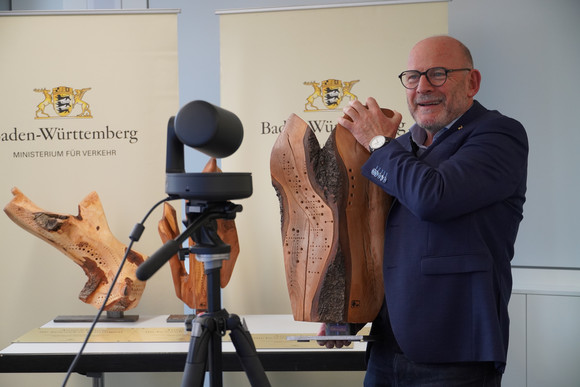 Minister Hermann steht vor einer Kamera und hält eine Holz-Skulptur in den Händen.