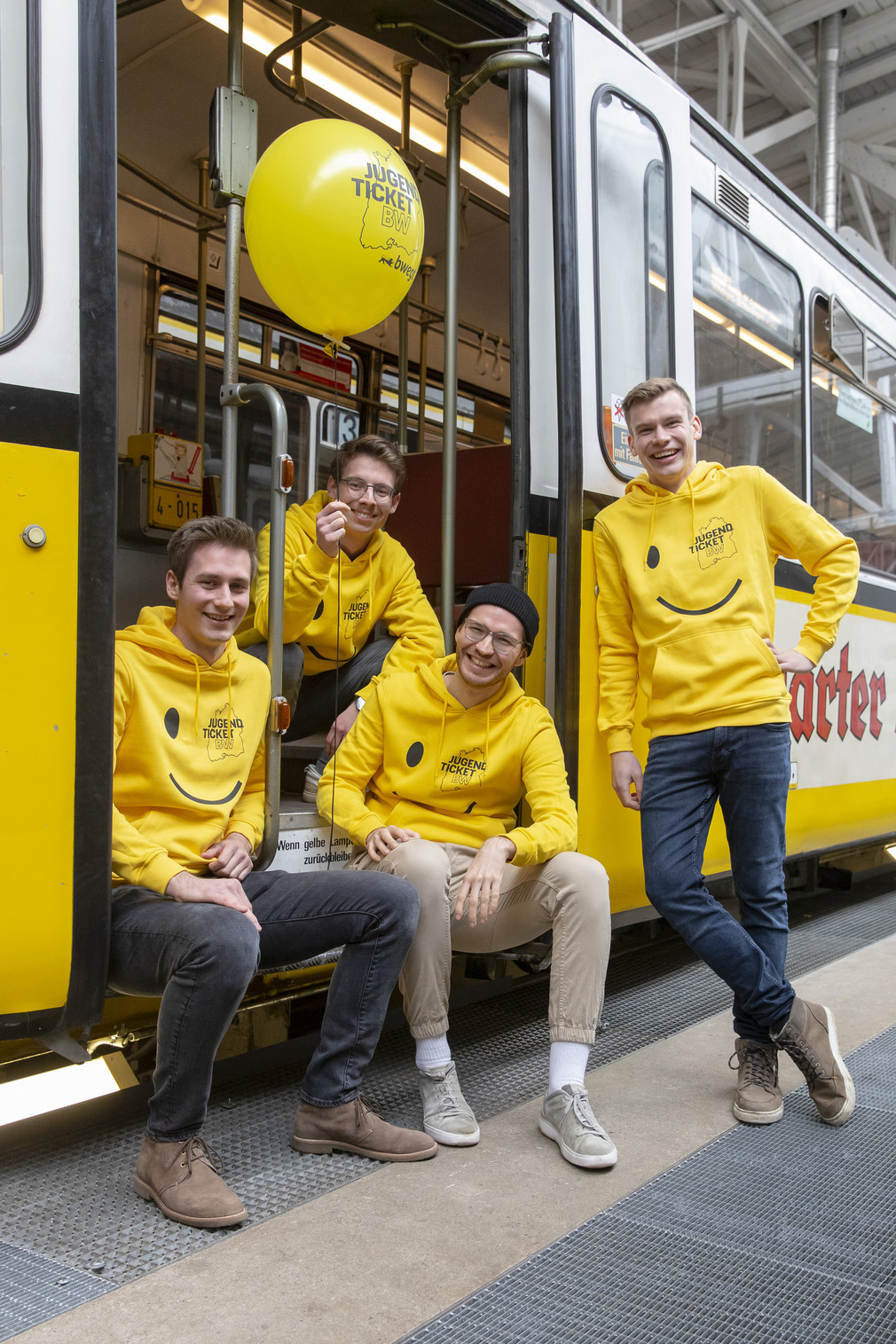 Vier fröhliche junge Männer in gelben Hoodies mit JugendticketBW-Design und mit einem ebensolchen Luftballon im Eingangsbereich einer alten Straßenbahn.