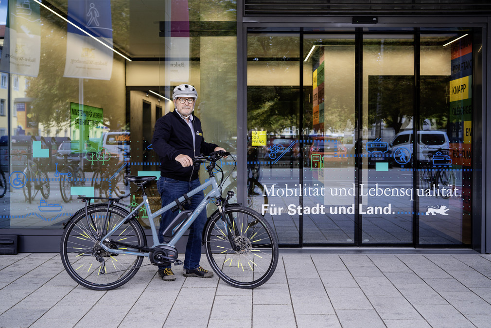 Verkehrsminister Winfried Hermann trägt einen Fahrradhelm und steht mit Fahrrad vor dem Eingang zum Verkehrsministerium.
