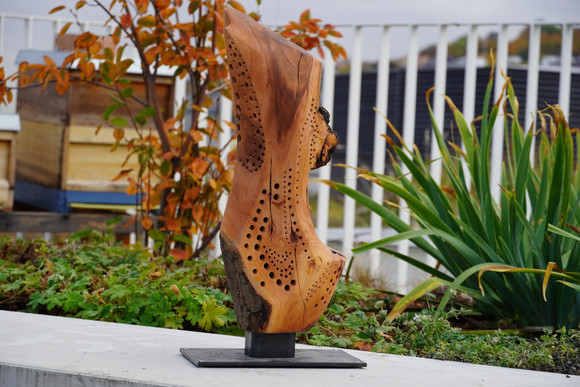 Holz-Skulptur auf Dachterrasse. Im Hintergrund sind Bienenkästen zu sehen.