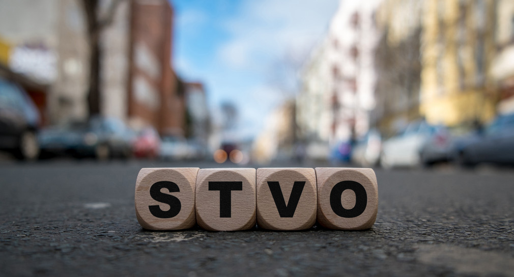 Auf einer Straße mit verschwommenem Hintergrund liegen vier Würfel mit jeweils einem Buchstaben. Dort steht: S – T – V – O.