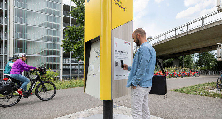 Ein Mann steht an einer Informationssäule und schaut auf sein Handy. Eine Radfahrerin fährt an ihm vorbei.