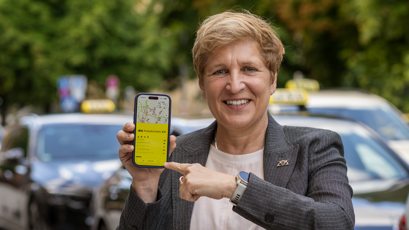 Verkehrsministerin Razavi steht neben Autos mit einem Handy in der Hand.