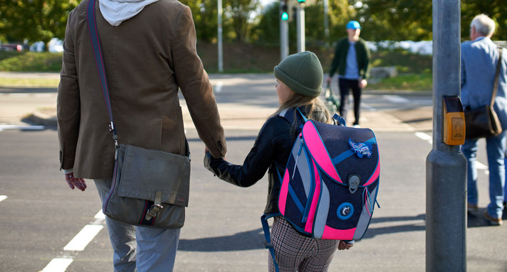Ein Mann und eine junge Schülerin stehen vor einer Ampel und halten Händchen.