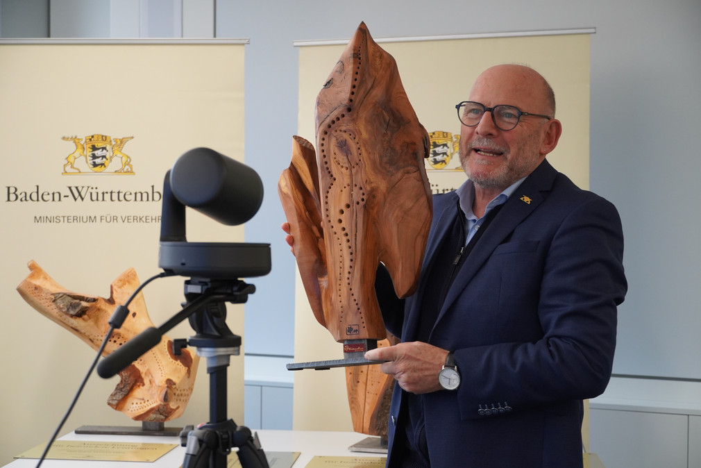 Minister Hermann steht vor einer Kamera und hält eine Holz-Skulptur in den Händen.