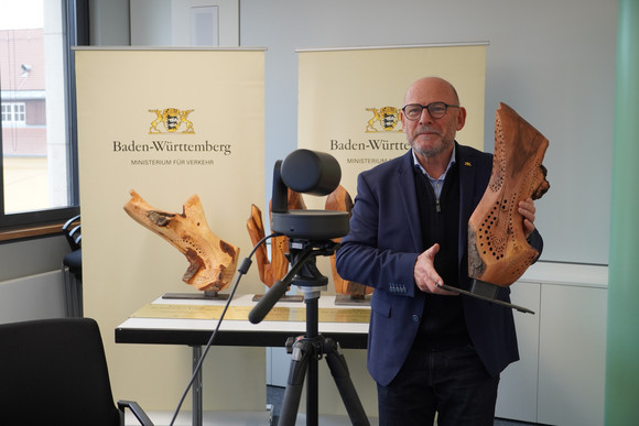 Minister Hermann steht vor einer Kamera und hält eine Holz-Skulptur in den Händen.