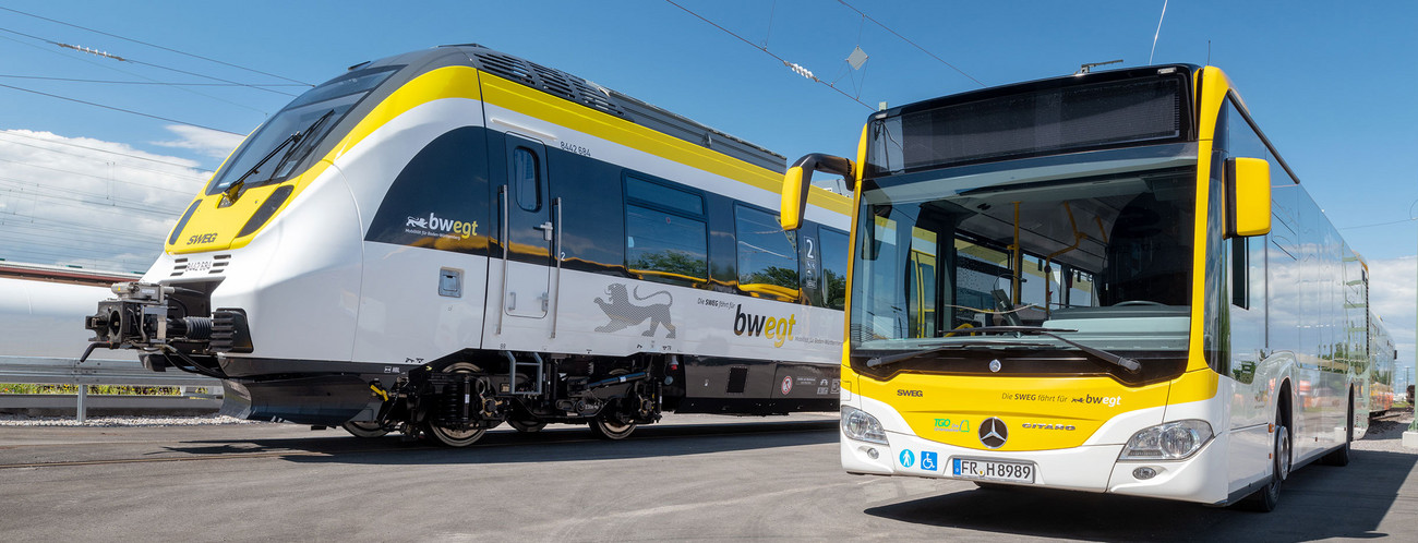 Ein Bus und zwei Züge stehen vor einer Werkshalle der SWEG nebeneinander.