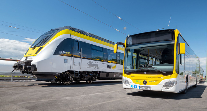 Ein Bus und zwei Züge stehen vor einer Werkshalle der SWEG nebeneinander.