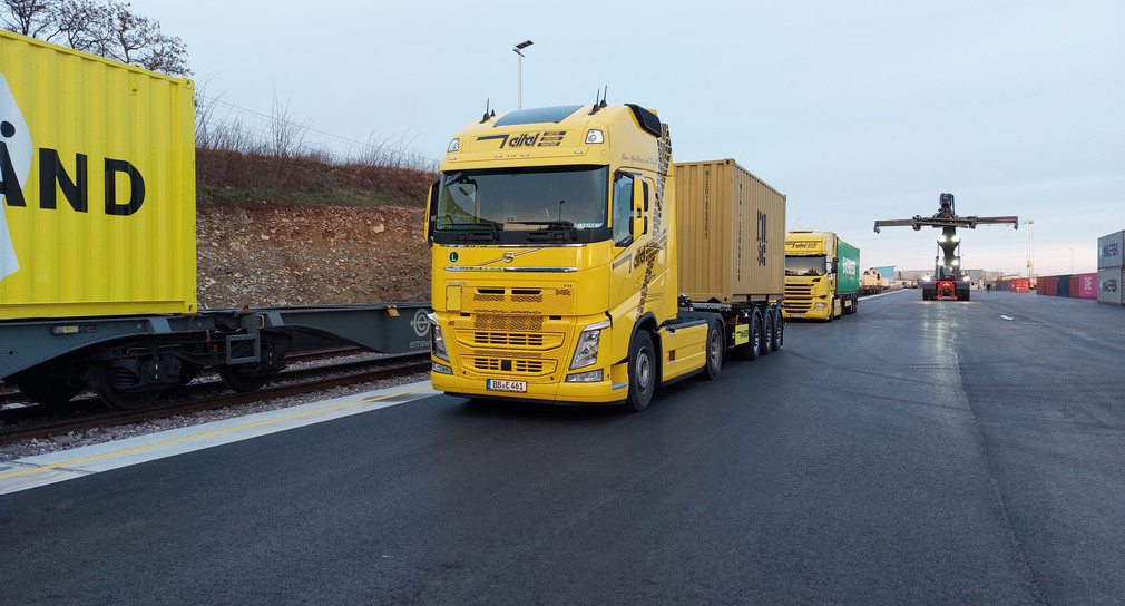 Lkw mit Container neben einem Güterzug mit THE-LÄND-Container.