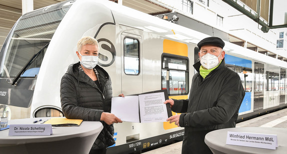 Dr. Nicola Schelling und Minister Hermann präsentieren den unterschriebenen Vertrag. Im Hintergrund sieht man eine Bahn.