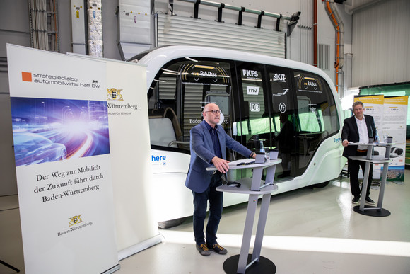 Verkehrsminister Winfried Hermann und Prof. Dr. Hans-Christian Reuss (FKFS) hinter ihren Rednerpults, im Hintergrund der autonome Shuttlebus 