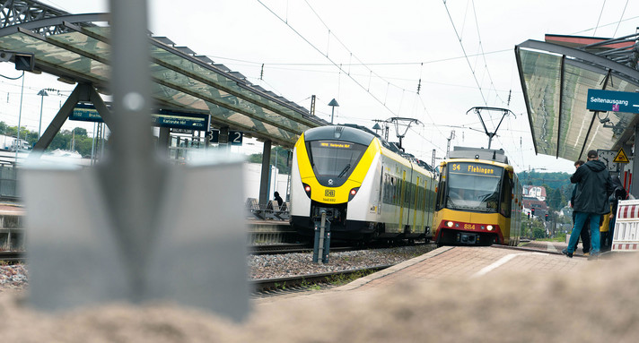 Zwei Züge stehen an einem Bahnsteig. Im Vordergrund steht ein Sandhaufen mit einem Spaten.