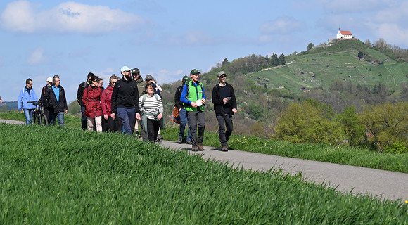 Wanderer laufen auf einem Weg und im Hintergrund sieht man eine Kapelle auf einem Berg