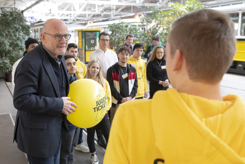 Minister Hermann hält einen gelben Luftballon in der Hand und spricht mit einer Person in gelbem Hoodie. Im Hintergrund steht eine Gruppe junger Menschen.
