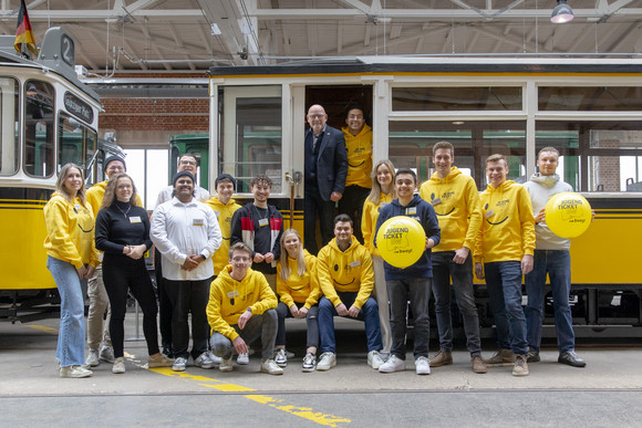 Minister Hermann im Eingangsbereich einer alten Straßenbahn, vor ihm eine Gruppe junger Menschen, teils mit gelben Hoodies im JugendticketBW-Design sowie mit ebensolchen Luftballons.