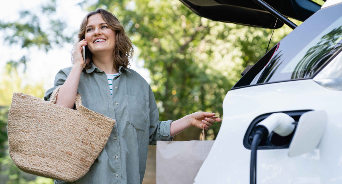 Frau mit Einkaufstasche neben einem ladenden Elektroauto.