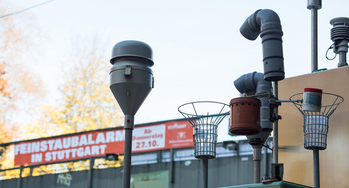 Ein Banner mit der Aufschrift "Feinstaubalarm", das Banner hängt an einer Fußgängerbrücke über der Bundesstraße 14 in Stuttgart. Im Vordergrund sind Messgeräte einer Feinstaubmessstation zu sehen.