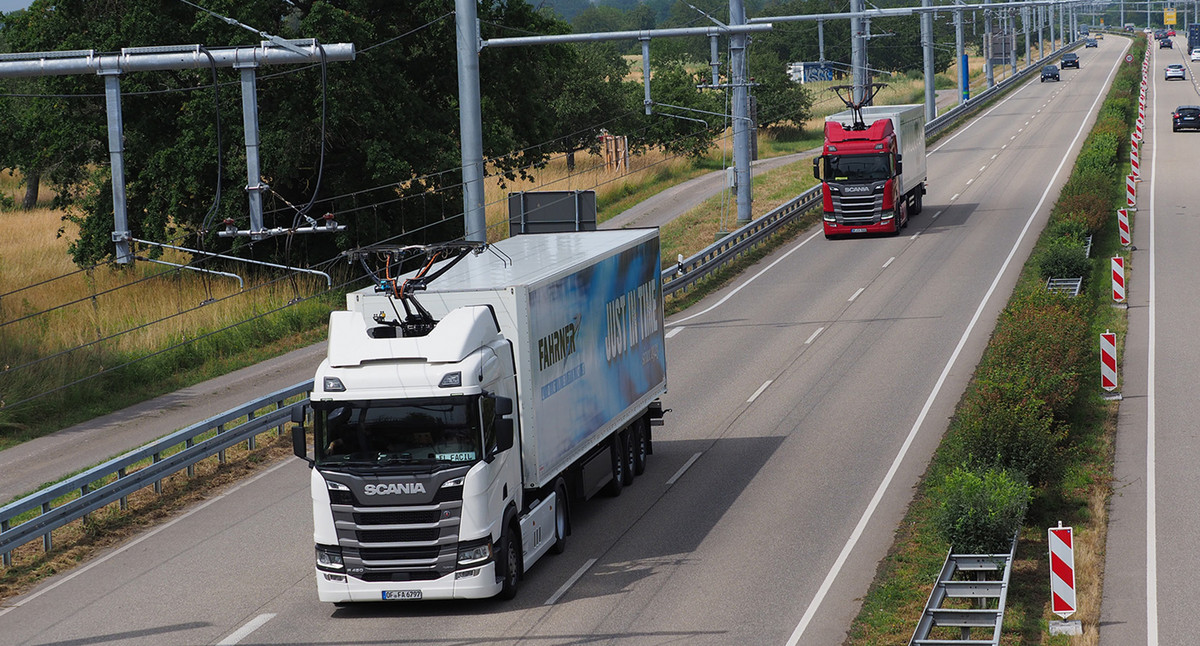 Zwei Lastwagen mit ausgeklapptem Stromabnehmer fahren hintereinander eine elektrifizierte Bundesstraße entlang.