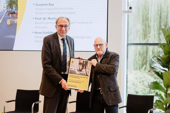 Verkehrsminister Winfried Hermann überreicht Prof. Dr. Martin Haag, Baubürgermeister der Stadt Freiburg, auf der Bühne einen symbolischen Förderbescheid für den LGVFG-Klimabonus für eine Maßnahme aus dem Freiburger Klimamobilitätsplan („Elsässer Straße: Umgestaltung des Straßenraums“)