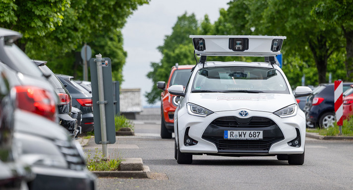 Ein Auto mit Kameras und Sensoren auf dem Dach fährt an Parkplätzen vorbei.
