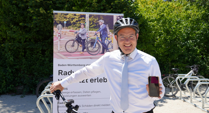 Staatssekretär Raimund Haser steht vor seinem Fahrrad mit einem Fahrradhelm auf dem Kopf und hält ein Handy in die Kamera.