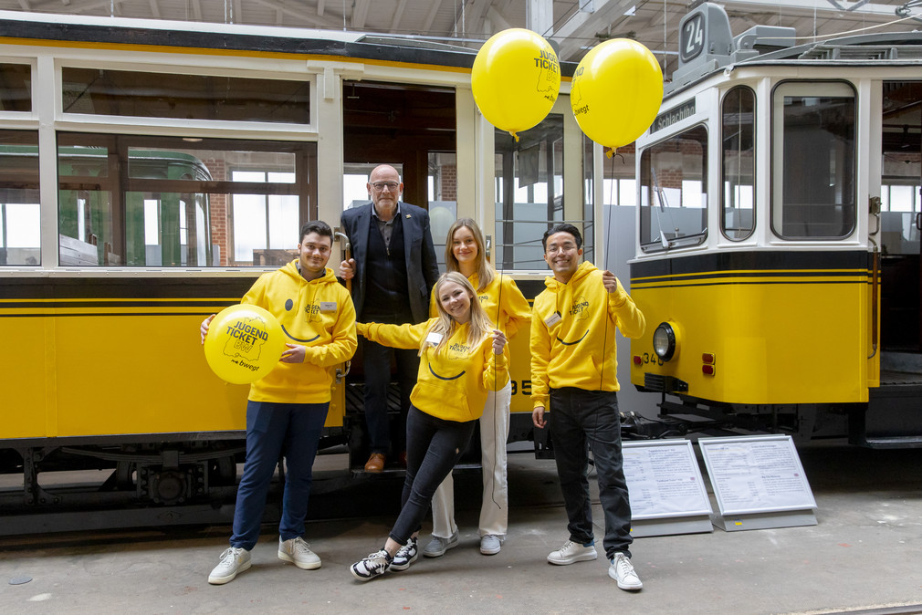 Vier fröhliche junge Menschen in gelben Hoodies mit JugendticketBW-Design und mit drei ebensolchen Luftballons vor einer alten Straßenbahn, während Minister Hermann im Eingangsbereich der Straßenbahn steht.