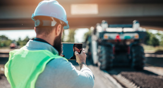 Ein Bauarbeiter steht auf einer Straße während im Hintergrund der Aspahlt neu gelegt wird. Er macht ein Foto.