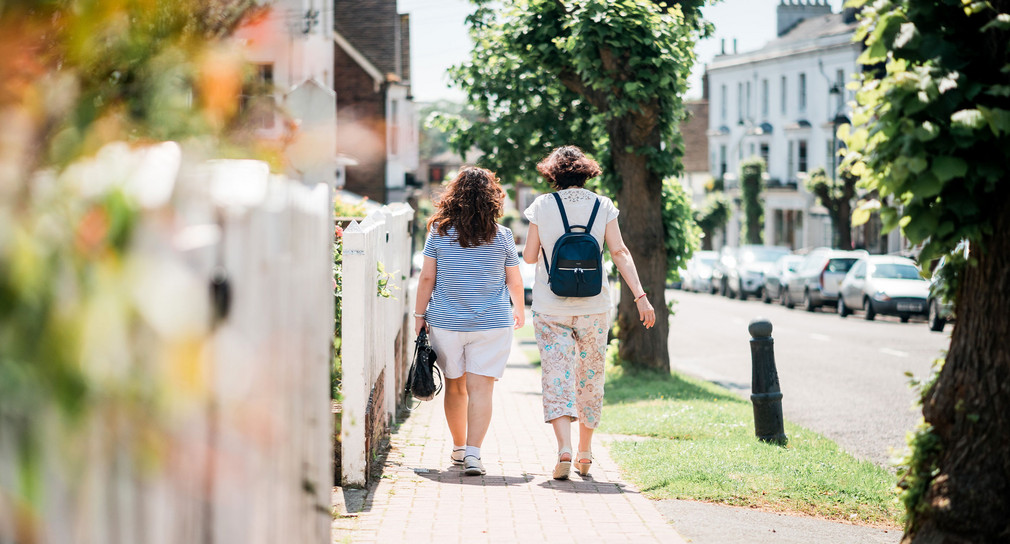 Zwei Frauen laufen nebeneinander auf einem Gehweg in einem grünen Wohngebiet