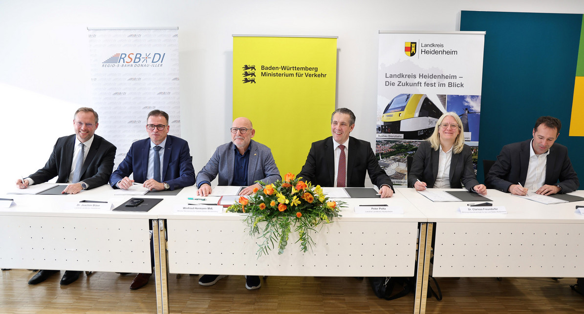 Eine Gruppe von Menschen sitzt am Tisch und schaut in die Kamera. Vor ihnen liegen Dokumente zum unterschreiben.