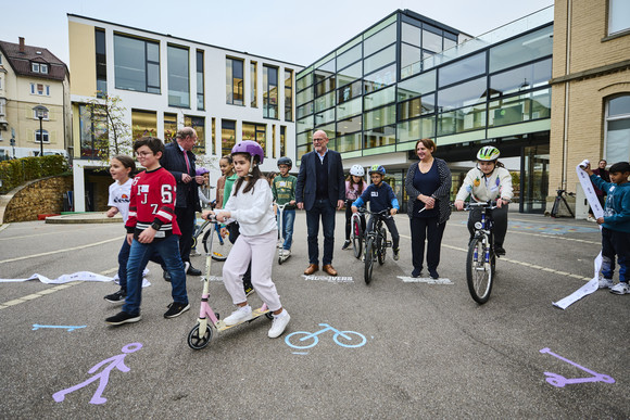 Verkehrssicherheit MOVERS – Aktiv zur Schule Minister Hermann, Ministerin Schopper und Innenstaatssekretär Klenk umgeben von Kindern zu Fuß, auf Fahrrädern und Rollern.