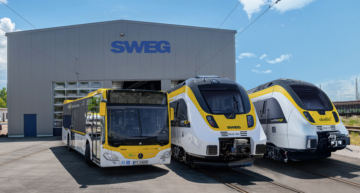 Ein Bus und zwei Züge stehen vor einer Werkshalle der SWEG nebeneinander.
