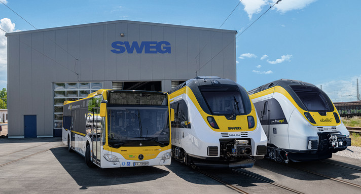 Ein Bus und zwei Züge stehen vor einer Werkshalle der SWEG nebeneinander.