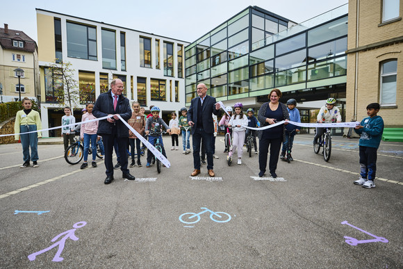 Verkehrssicherheit MOVERS – Aktiv zur Schule Minister Hermann, Ministerin Schopper und Innenstaatssekretär Klenk beim Startschuss von MOVERS, während sie das Band durchschneiden. Im Hintergrund Kinder zu Fuß, mit dem Fahrrad oder Roller.
