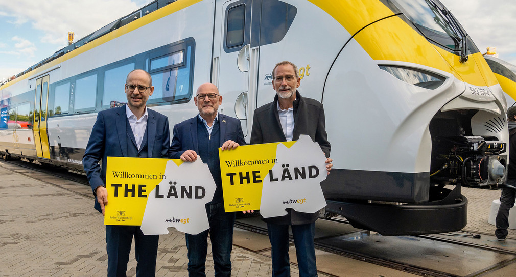 Gerhard Greiter, Winfried Hermann und Volker M. Heepen stehen vor dem neuen Mireo Zug von Siemens und halten jeweils ein Schild "Welcome to the Länd" in der Hand.