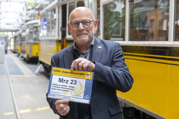 Minister Hermann mit einem naldo-JugendticketBW in der Hand, im Hintergrund alte Straßenbahnen.