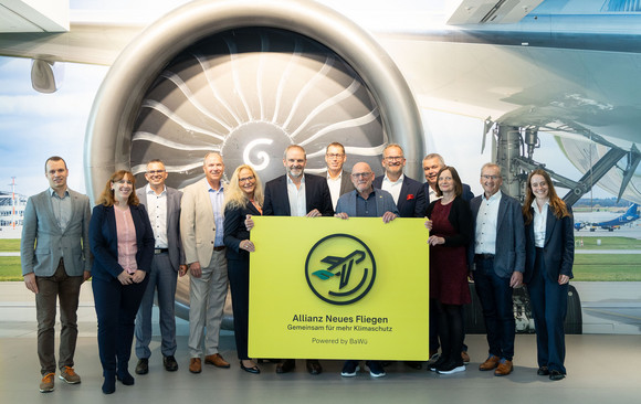 Menschen stehen zu einem Gruppenbild zusammen. Im Hintergrund ist ein Foto von einem Flugzeugtriebwerk abgebildet.