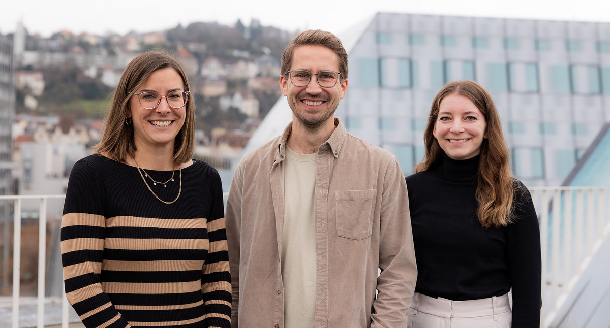 Zwei Frauen und ein Mann stehen auf einer Dachterrasse mit Stuttgart im Hintergrund.