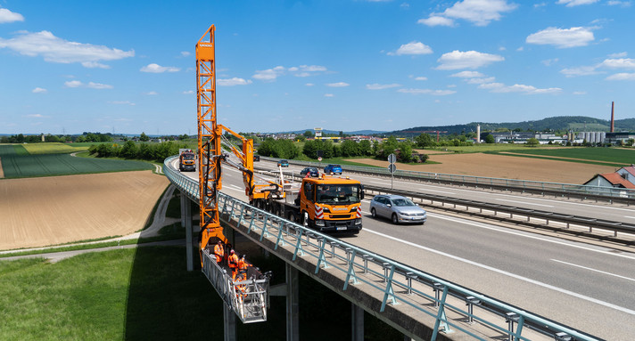 Analyse Zustand der Brücken in Baden-Württemberg Eine mehrspurige Straßenbrücke führt über ein Tal. Ein Lastwagen mit einer ausfahrbaren Brücke bringt Bauarbeiter unter die Fahrbahn, um die Unterseite der Brücke zu begutachten.