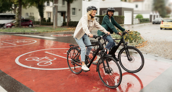 Zwei Radfahrerinnen überqueren auf einem rot markierten Radweg eine Straße.