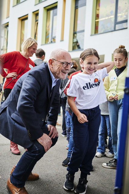 Verkehrssicherheit MOVERS – Aktiv zur Schule Minister Hermann, halb in der Hocke, einem Mädchen zugewandt, schaut sich mit diesem zusammen etwas an. Beide lachen. Im Hintergrund weitere Erwachsene und Kinder.