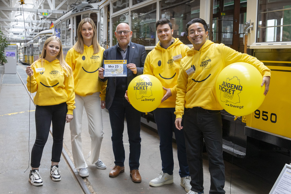 Vier fröhliche junge Menschen in gelben Hoodies mit JugendticketBW-Design und mit zwei ebensolchen Luftballons. Zwischen Ihnen Minister Hermann. Im Hintergrund alte Straßenbahnen.