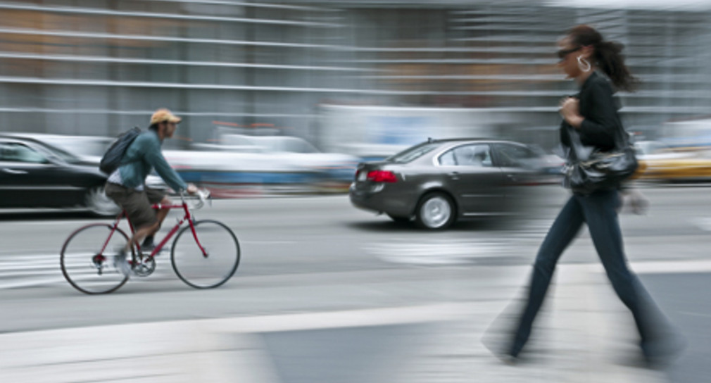 Radfahrer fährt auf Straße zwischen Autos. Auf dem Gehweg läuft eine Fußgängerin.