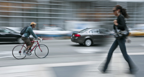Radfahrer fährt auf Straße zwischen Autos. Auf dem Gehweg läuft eine Fußgängerin.