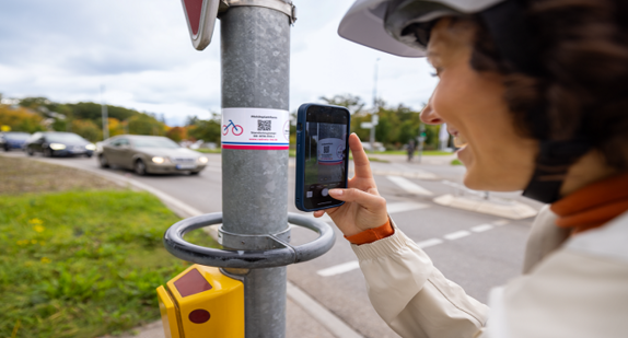 Eine Frau steht an einer Ampel und scannt mit ihrem Handy einen QR-Code von einem Aufkleber der Radkultur ab. Der QR-Code führt sie direkt zum RadMELDER.